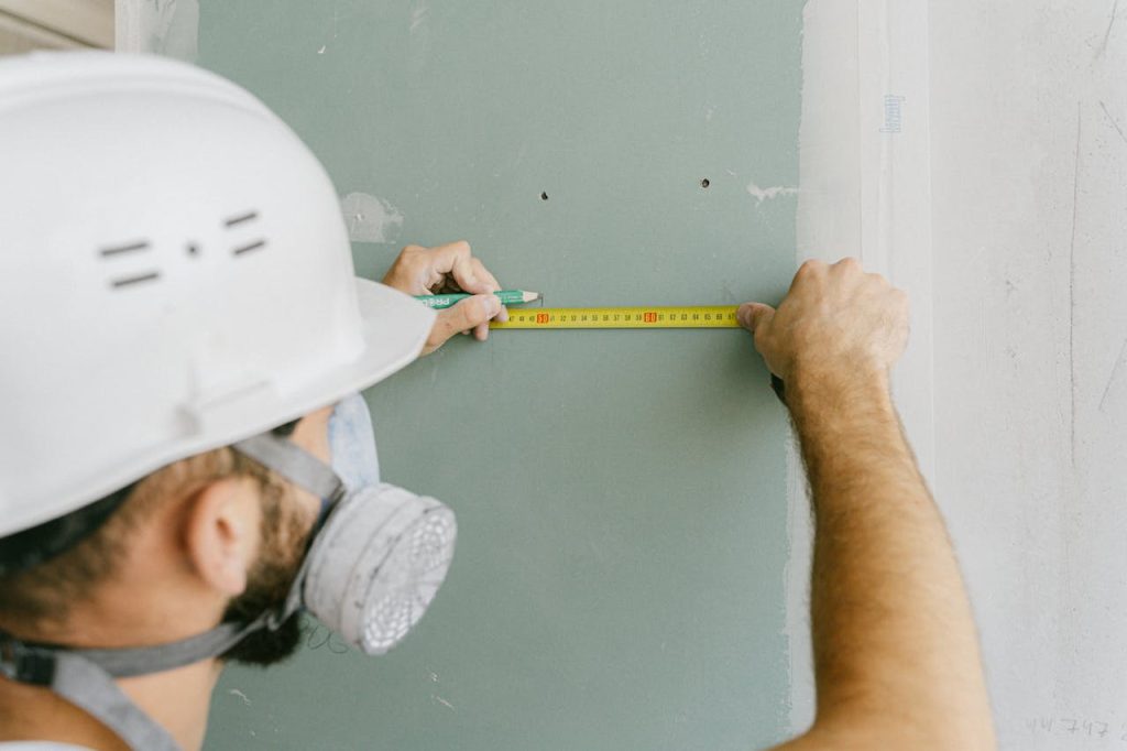 person-holding-a-tape-measure-5493663 A construction worker precisely measuring drywall, wearing a helmet and mask for safety.